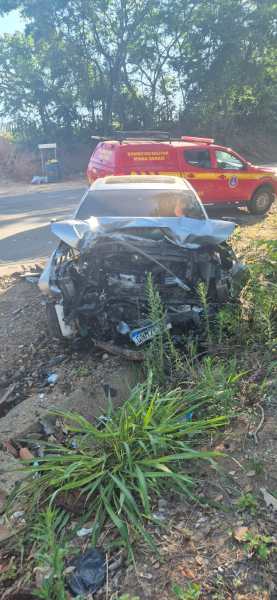 Carro acidentado na beira da estrada, com a parte da frente danificada e gramado ao redor. Ao fundo, uma viatura de emergência está estacionada.
