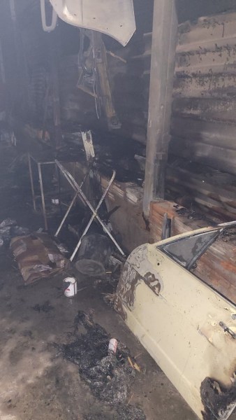 Imagem de um espaço danificado por incêndio, mostrando um carro parcialmente queimado, restos de móveis e objetos carbonizados em um ambiente escuro e em desordem.