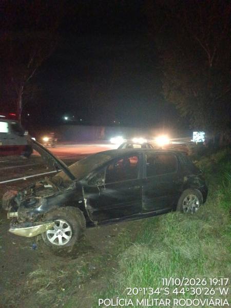 Veículo acidentado à noite, com o capô levantado, na beira da estrada, cercado por árvores.