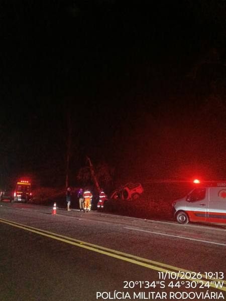 Cena de um acidente de carro à noite, com veículos de emergência e socorristas no local, próximo a um carro capotado na margem da estrada.