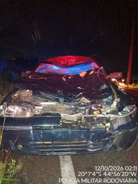 Frente de um carro danificado após um acidente, com o capô amassado e luzes azuis piscando ao fundo em um ambiente noturno.