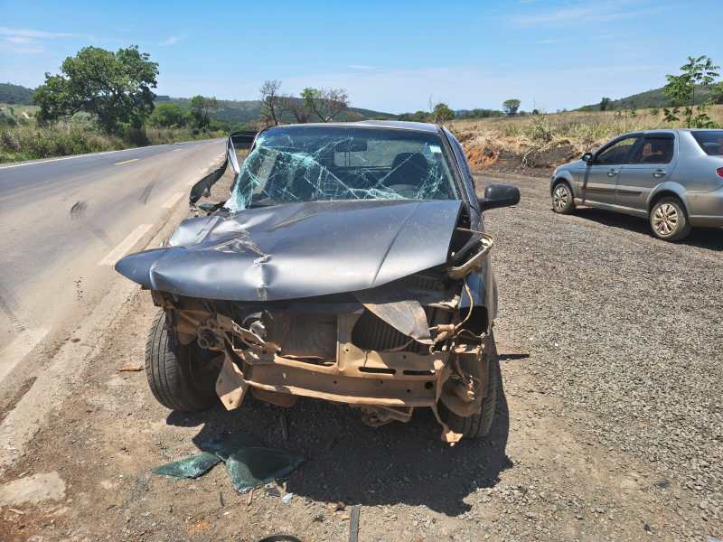 Veículo acidentado com a parte frontal danificada em uma estrada rural, com para-brisa quebrado e peças de vidro no chão.