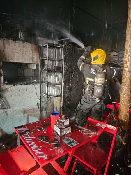 Bombeiro combatendo incêndio em um local danificado, com paredes e prateleiras negras de fumaça, e mesas vermelhas de plástico.