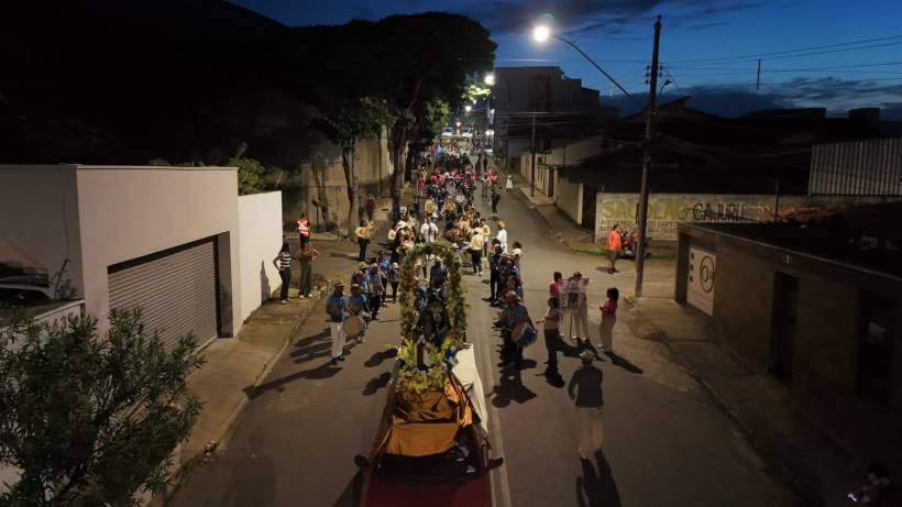 Uma procissão noturna em uma rua com iluminação, onde um grupo de pessoas caminha em direção a um carro alegórico decorado com flores.