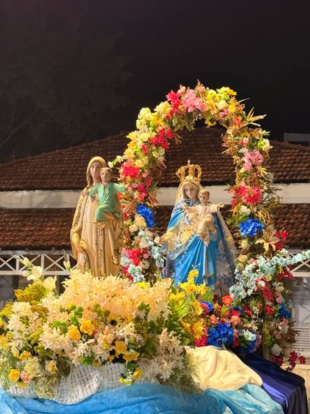 Estátua da Virgem Maria com flores ao redor, acompanhada por figuras de São José e do menino Jesus, durante uma celebração noturna.