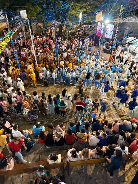 Vista aérea de um evento festivo com grande multidão reunida, destacando pessoas em trajes coloridos e decorativos, com uma estrutura de palco ao fundo e decoração em azul.