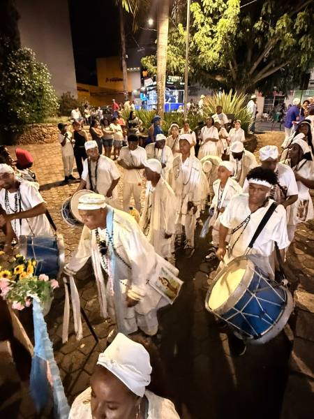Grupo de pessoas em trajes brancos tocando tambores durante uma celebração noturna, rodeados por árvores e iluminação urbana.