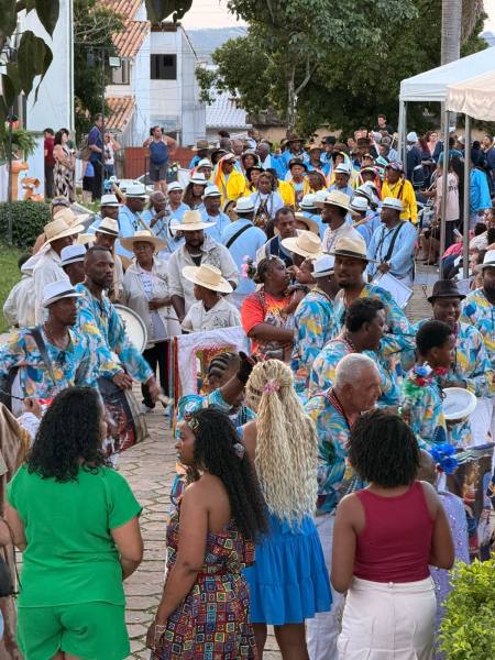 Multidão em uma celebração cultural, com grupos de músicos e dançarinos usando roupas coloridas e chapéus, enquanto pessoas assistem ao evento.