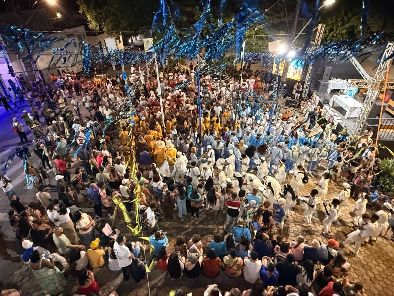 Vista aérea de uma grande festa ao ar livre, com multidão dançando e celebrando sob decorações coloridas. Pessoal vestido em trajes variados, com um palco visível ao fundo.