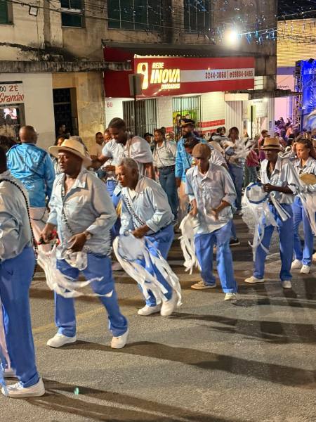 Grupo de pessoas dançando em um desfile, vestindo roupas azuis e brancas, com alguns usando chapéus, durante um evento festivo à noite.