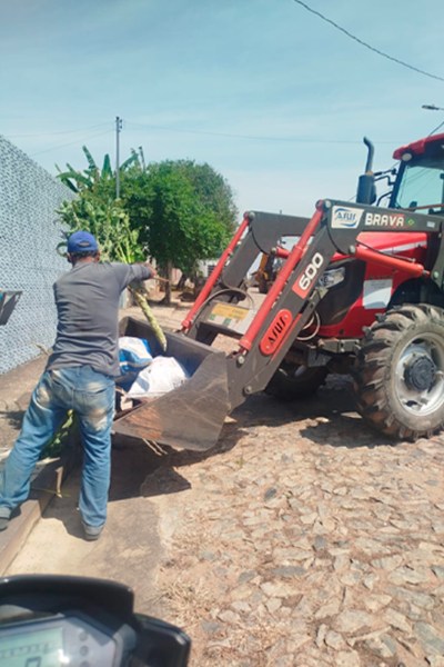 Homem operando uma pá carregadeira vermelha em uma rua pavimentada, levantando uma carga de sacos e plantas.