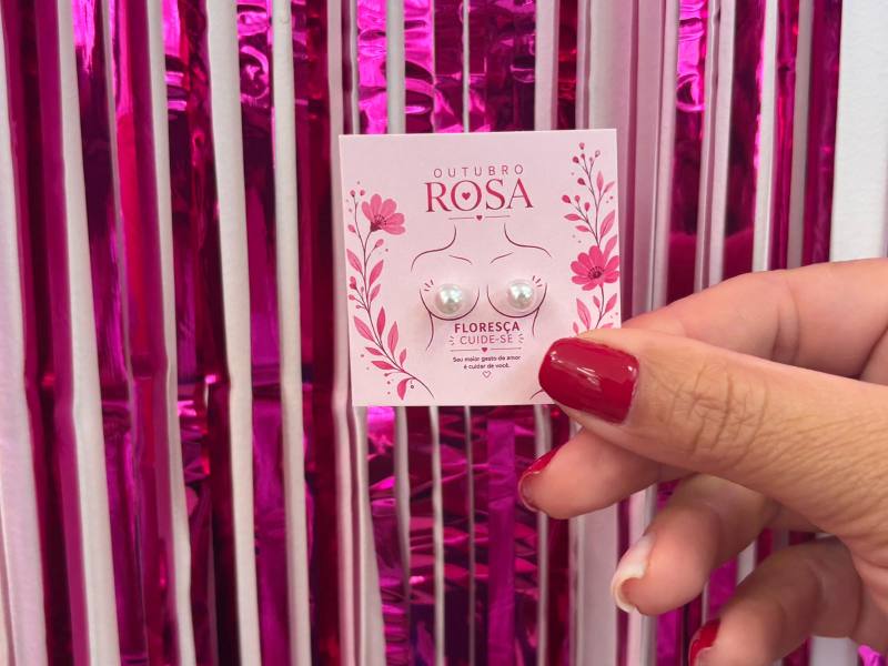 Imagem de um cartão com brincos em forma de pérolas, decorado com a frase 'Outubro Rosa' e elementos florais, segurado por uma mão com unhas vermelhas, em frente a uma parede rosa.