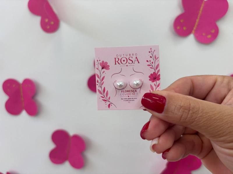Mão segurando um cartão rosa com dois brincos em formato de pérola, com fundo de borboletas rosas.
