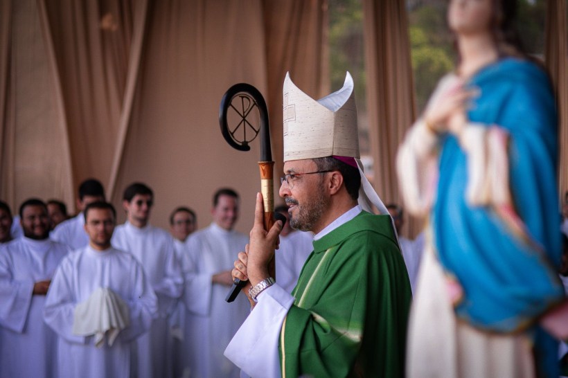 Um bispo em traje litúrgico verde, com um chapéu pontifício, segurando um bastão enquanto faz um discurso, ao fundo, várias pessoas vestindo roupas brancas e uma estátua de uma santidade.