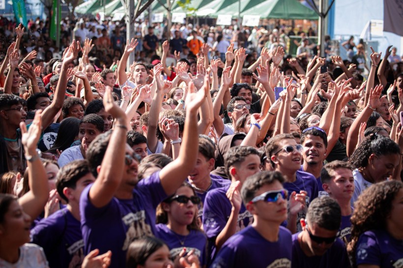 Um grande público em um evento ao ar livre, com pessoas radiantes levantando as mãos e celebrando. Muitos usam camisetas roxas e parecem estar se divertindo.