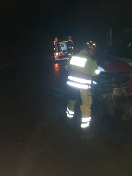 Um bombeiro com uniforme refletivo está trabalhando em um veículo, enquanto um caminhão de bombeiros aparece ao fundo em uma cena noturna.