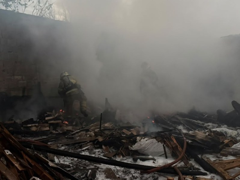 Bombeiros atuando em área coberta de fumaça após um incêndio, com escombros de madeira visíveis.