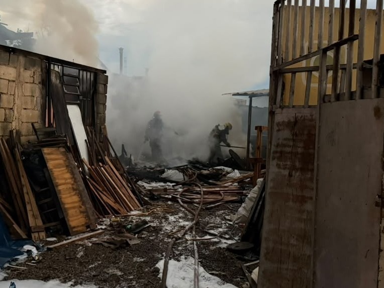 Bombeiros combatendo incêndio em área coberta de fumaça, com detritos de madeira ao redor.