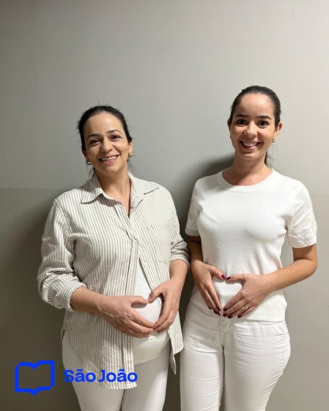 Duas mulheres grávidas posando e fazendo um coração com as mãos na barriga, em um ambiente neutro.