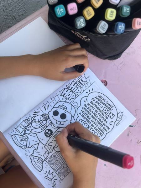 Uma criança colorindo um desenho em um caderno com uma caneta, com uma ilustração de um personagem de hip hop.