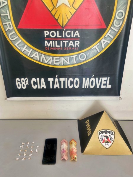 Imagem de uma mesa com uma bandeira da Polícia Militar de Minas Gerais ao fundo, um celular, dois pacotes de drogas e uma pirâmide de identificação da PMMG.