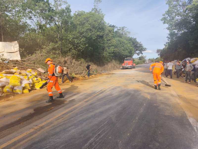 Equipes de trabalhadores em trajes de segurança laranja realizando limpeza na estrada, com sacos amarelados ao fundo. Um caminhão é visível na estrada ao fundo.