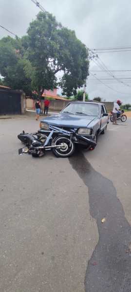 Um carro azul colidido com uma motocicleta caída na rua, com pessoas ao fundo observando a cena.
