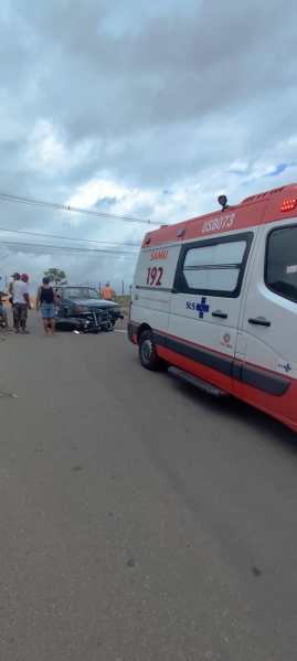 Ambulância parada em uma rua com um carro acidentado e pessoas ao redor.