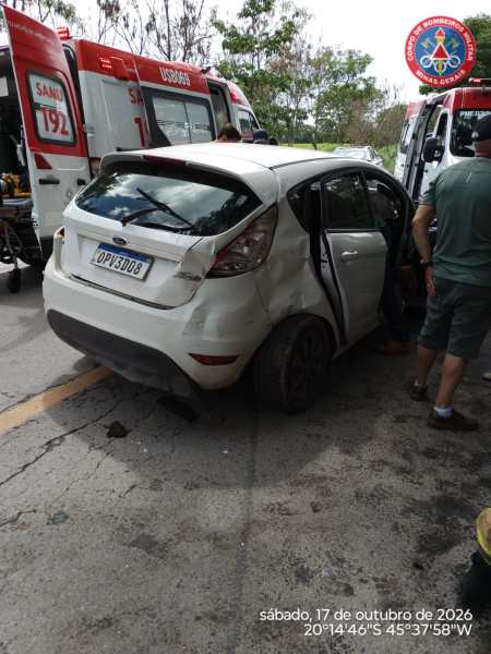 Veículo branco danificado em acidente, com a porta traseira aberta, próximo a ambulâncias e pessoas ajudando na cena.