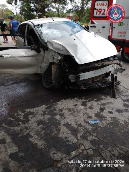 Carro branco danificado na parte frontal após um acidente, com pessoas ao fundo e veículo de emergência visível.