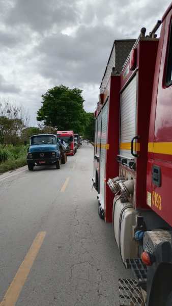 Vista de um caminhão de bombeiros estacionado à beira da estrada, com um pouco de vegetação ao fundo e outros veículos alinhados na via.