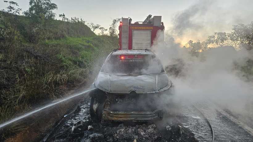 Carro queimado sendo resfriado pelos bombeiros ao fundo, com fumaça saindo do veículo em uma estrada.