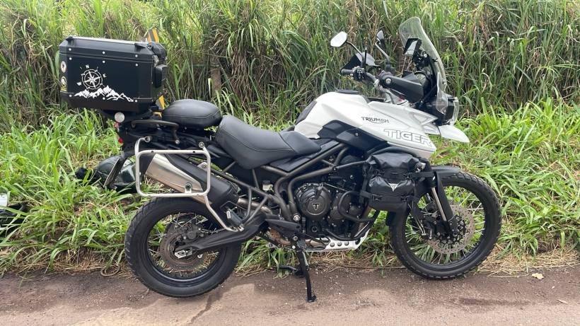 Motocicleta Triumph Tiger 800 estacionada ao lado de grama alta, equipada com um baú preto na parte traseira.