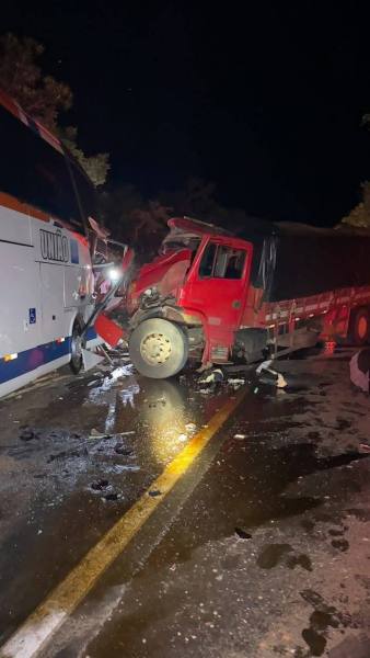 Acidente noturno envolvendo um caminhão vermelho e um ônibus, com danos visíveis na parte frontal do caminhão e sujeira na pista.