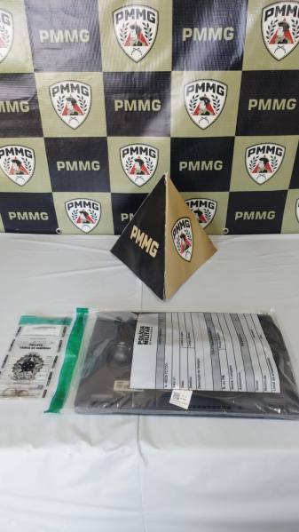 Piramide decorativa com o logotipo da PMMG ao lado de um pacote e documentos em uma mesa, com fundo de banners da PMMG.