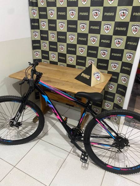 Uma bicicleta mountain bike colorida em frente a uma parede com logo PMMG e uma mesa ao fundo.