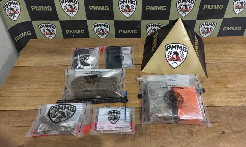 Imagem mostrando pacotes de substâncias não identificadas, organizados sobre uma mesa de madeira, com um fundo estampado com o logo da PMMG.