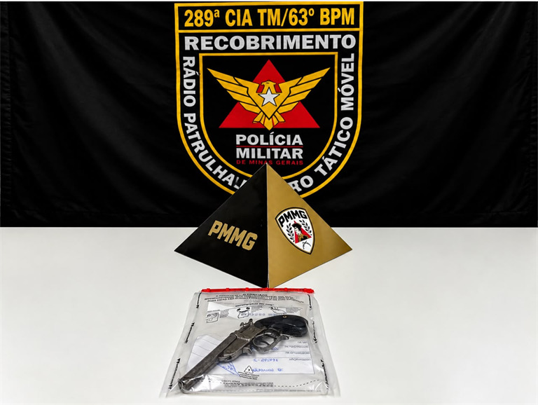 Imagem de objetos relacionados à polícia militar, incluindo um banner com a insígnia da Polícia Militar de Minas Gerais, dois pirâmides de identificação e uma arma em um saco transparente sobre uma mesa.
