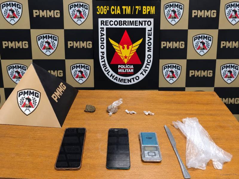 Imagem de uma mesa com dois celulares, uma balança, um canivete, e vários pacotes de substâncias aparentando ser drogas, com um banner da Polícia Militar ao fundo.