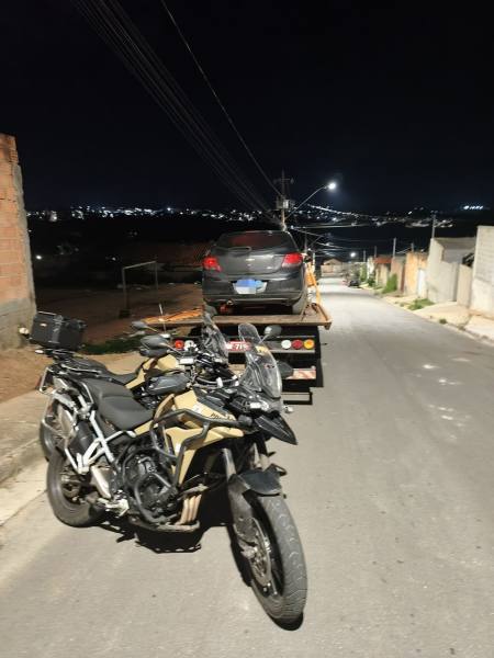 Motocicleta em primeiro plano com caminhão guincho carregando um carro ao fundo, em uma rua à noite.