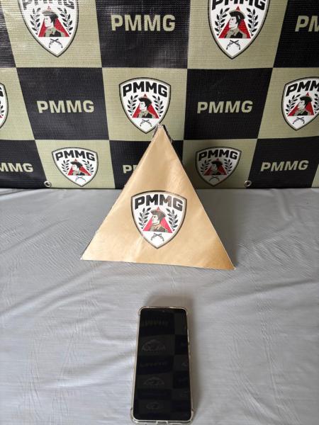 Piramide dourada com o emblema PMMG na frente, ao lado de um celular em uma mesa com fundo quadriculado de logotipos PMMG.
