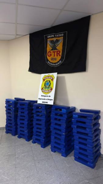 Imagem de uma parede com uma bandeira do Grupo Tático Rodoviário (GTR) e uma placa da Polícia Federal, ao lado de pilhas de caixas azuis empilhadas.