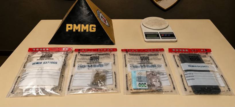 Pacotes lacrados com evidências, incluindo substâncias e dinheiro, em uma mesa com balança ao fundo e um troféu da PMMG.