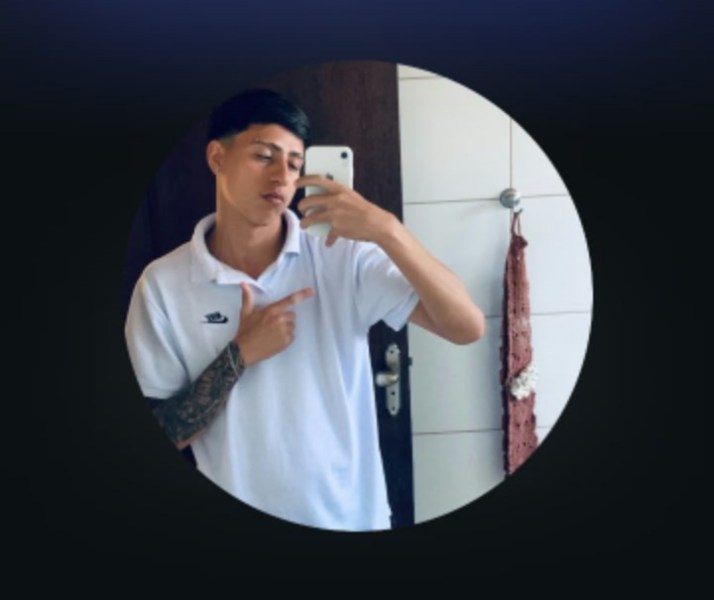 Jovem posa para uma selfie em um espelho, vestindo uma camiseta branca e com um tatuagem no braço esquerdo.