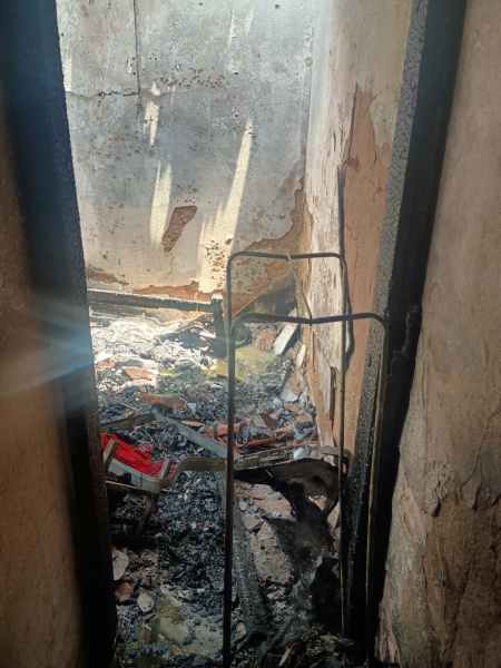 Vista de um espaço interno danificado por incêndio, com paredes parcialmente destruídas e restos de materiais queimados no chão.