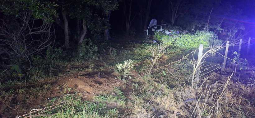 Cena noturna de um local com vegetação densa, mostrando vestígios de um acidente de carro com um veículo visível entre as árvores.