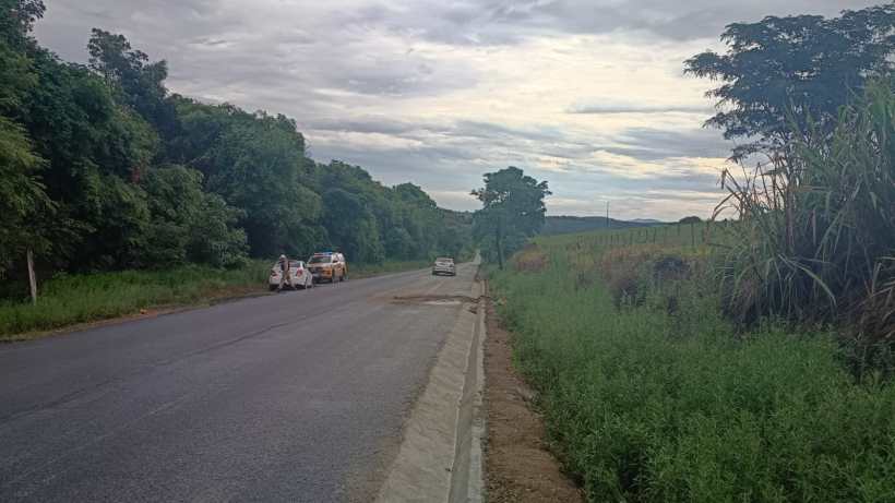 Uma estrada rural com vegetação densa ao lado, carros estacionados e um céu nublado ao fundo.