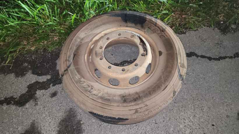 Roda de carro suja e desgastada, com buracos visíveis, posicionada sobre uma superfície de asfalto ao lado de grama.