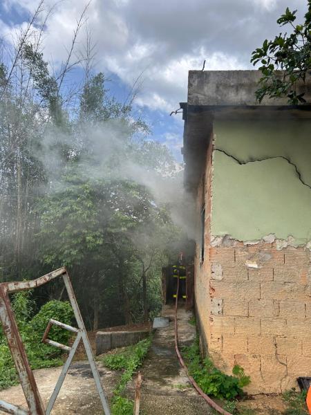 Cenário de incêndio em uma casa, com fumaça saindo da parte traseira e um caminhão de bombeiros visível ao fundo, cercado por vegetação.