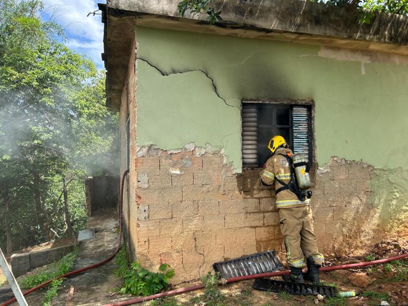 Bombeiro em frente a uma casa danificada, com fumaça saindo e uma parede quebrada, tentando acessar pela janela.